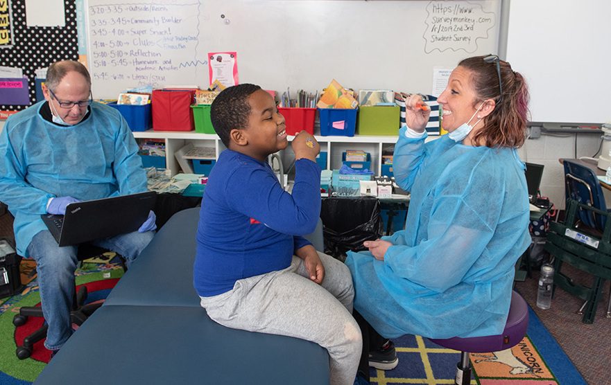 Dental provider and child practicing brushing teeth together in a classroom.