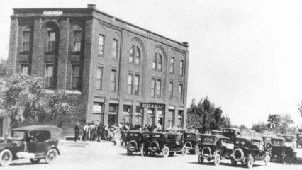 Black and white photo of several vintage cars parked in front of a brick building with a sign reading "Hotel." Several people are gathered outside the entrance.