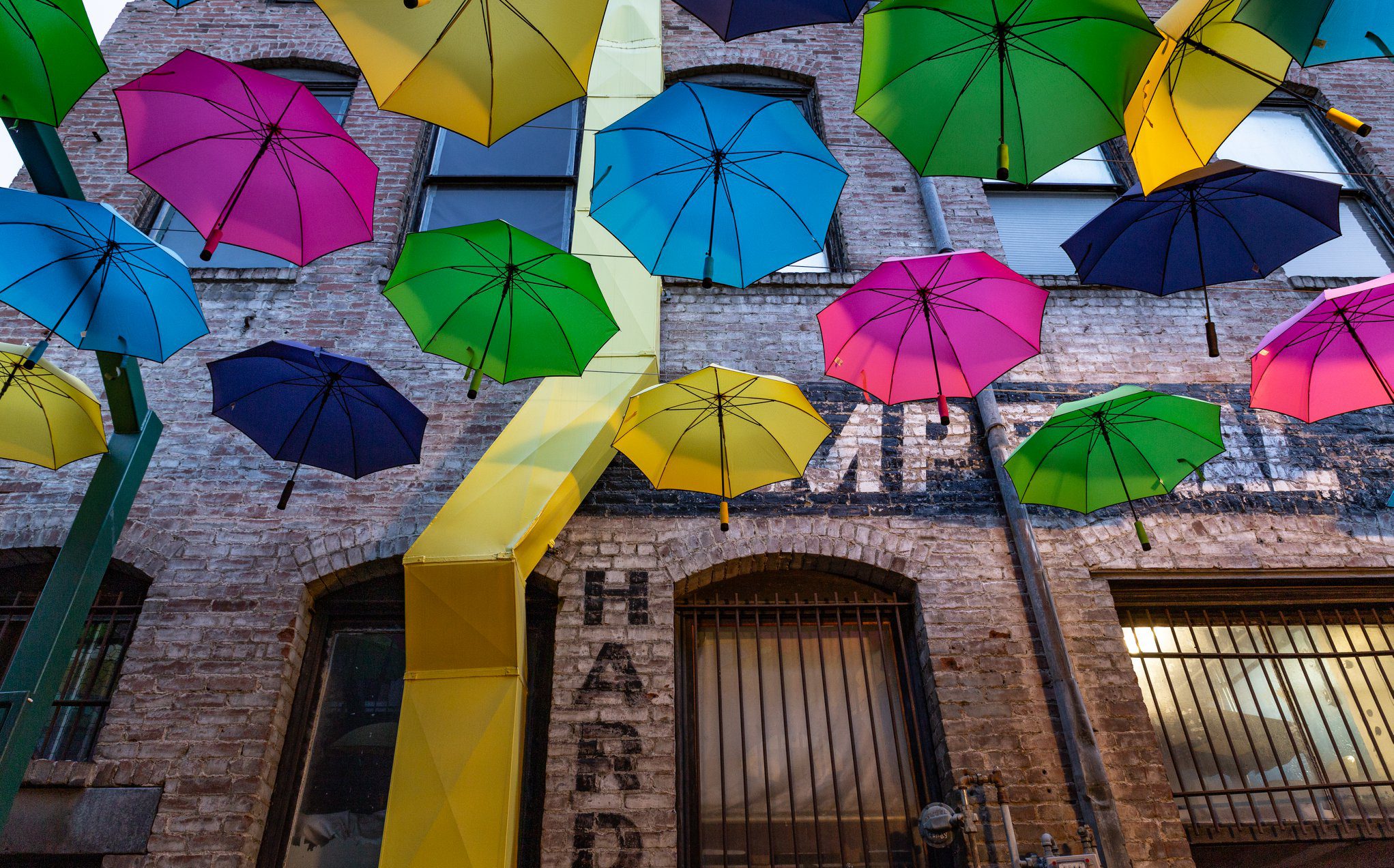 An alley featuring pink, blue, yellow, and green umbrellas with a brick building in the background.