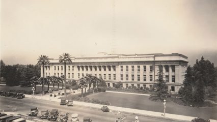 A historic building with large columns surrounded by tall trees and grass. Several vintage cars are parked along the street in front.
