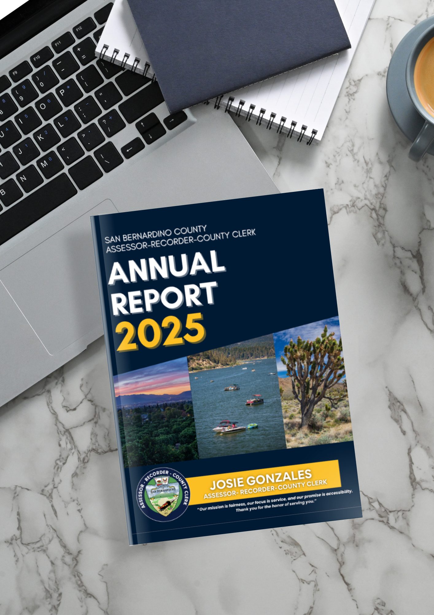 A laptop, notebooks, cup of coffee, and San Bernardino County Assessor-Recorder-County Clerk Josie Gonzales’s 2025 annual report on a marble table.