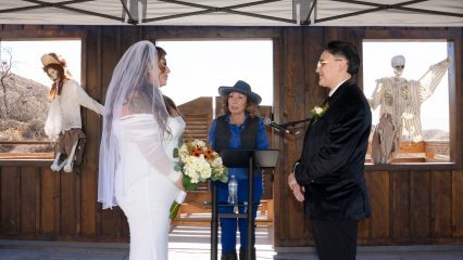 A wedding ceremony set in a rustic outdoor venue with wooden walls and open windows. Two people stand facing each other; one is holding a bouquet of flowers. A person officiates the ceremony using a microphone. In the background, decorative skeletons are visible.