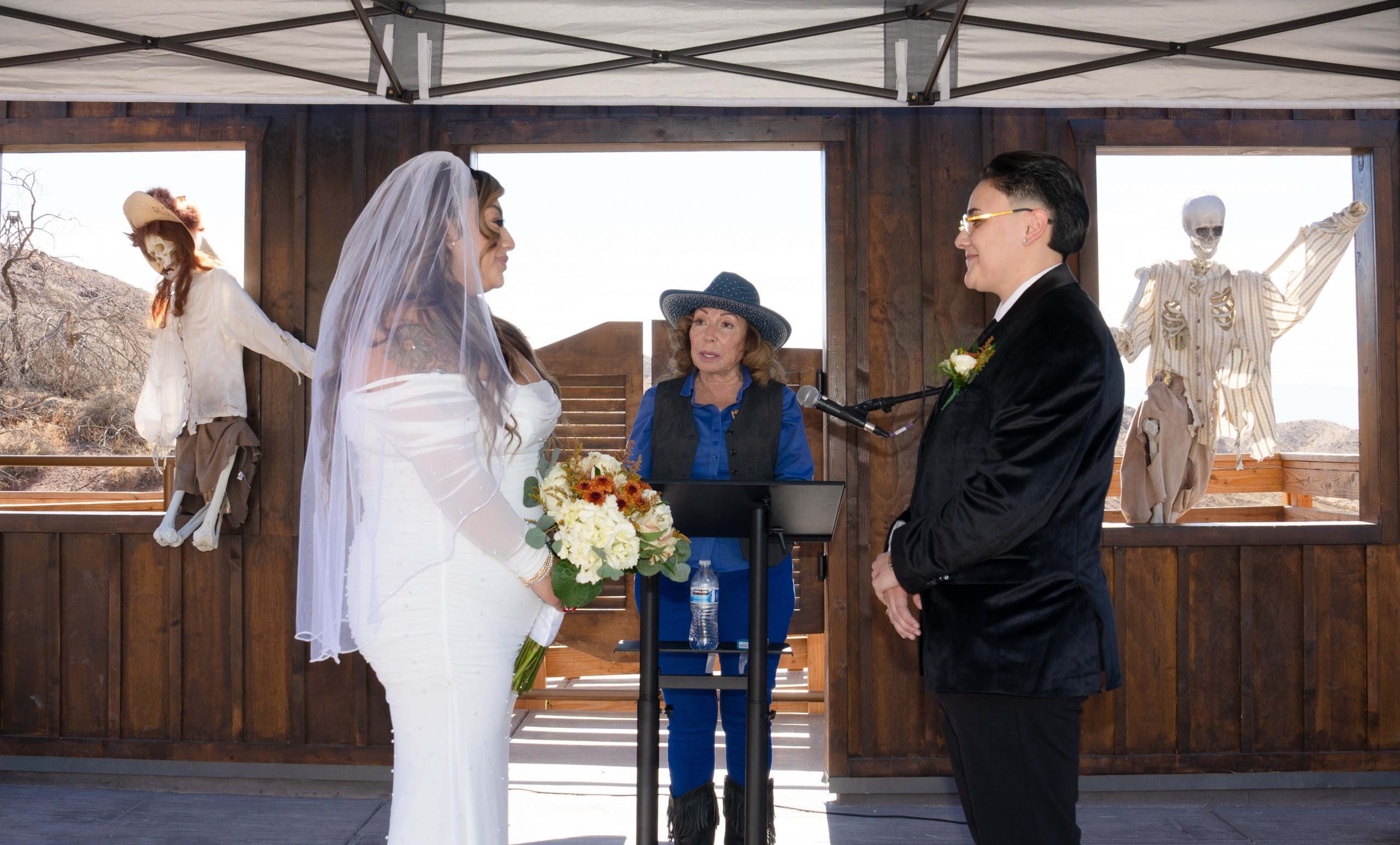 A wedding ceremony set in a rustic outdoor venue with wooden walls and open windows. Two people stand facing each other; one is holding a bouquet of flowers. A person officiates the ceremony using a microphone. In the background, decorative skeletons are visible.