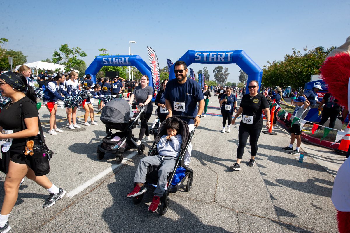People participate in a lively outdoor race, with strollers passing a blue start line. Cheerleaders and banners add a festive atmosphere.