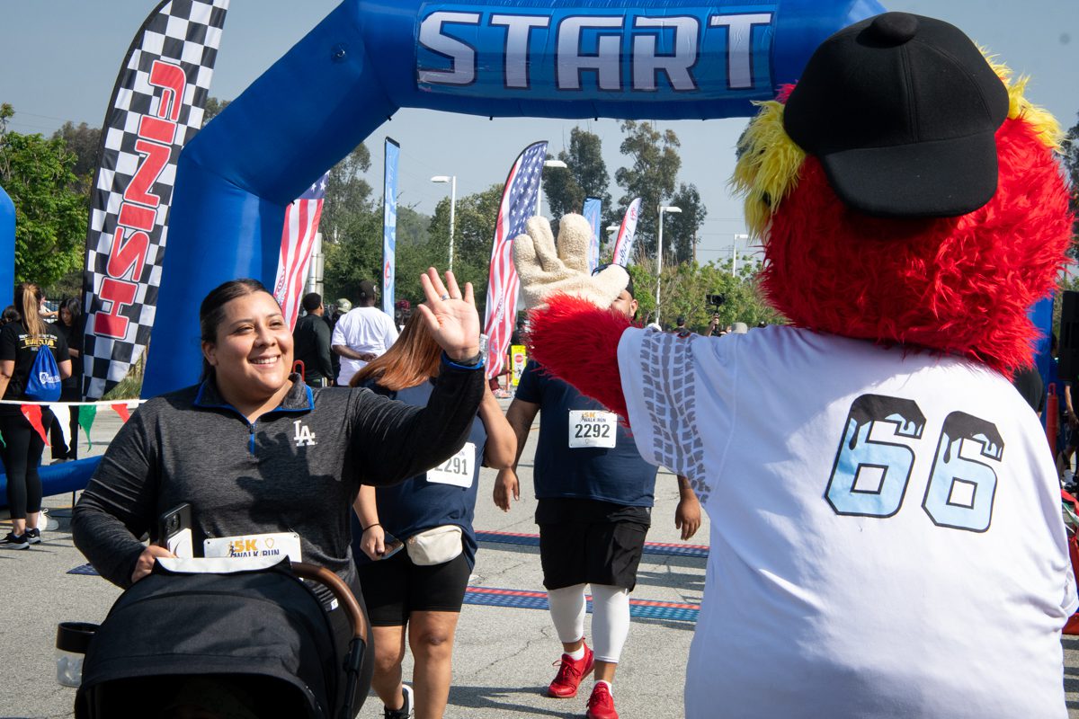 Woman giving a high-five to a mascot in a baseball cap at the start of a race. She smiles, holding a stroller. Participants and "START" arch in view. Energetic mood.