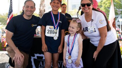 A smiling group of two adults and two children posing with medals in front of a podium at an outdoor event. The atmosphere is joyful and celebratory.