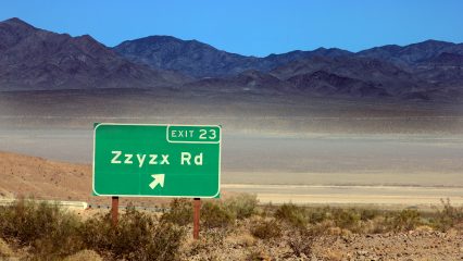 Road sign indicating Exit 23 for Zzyzx Road with a desert landscape and mountains in the background.