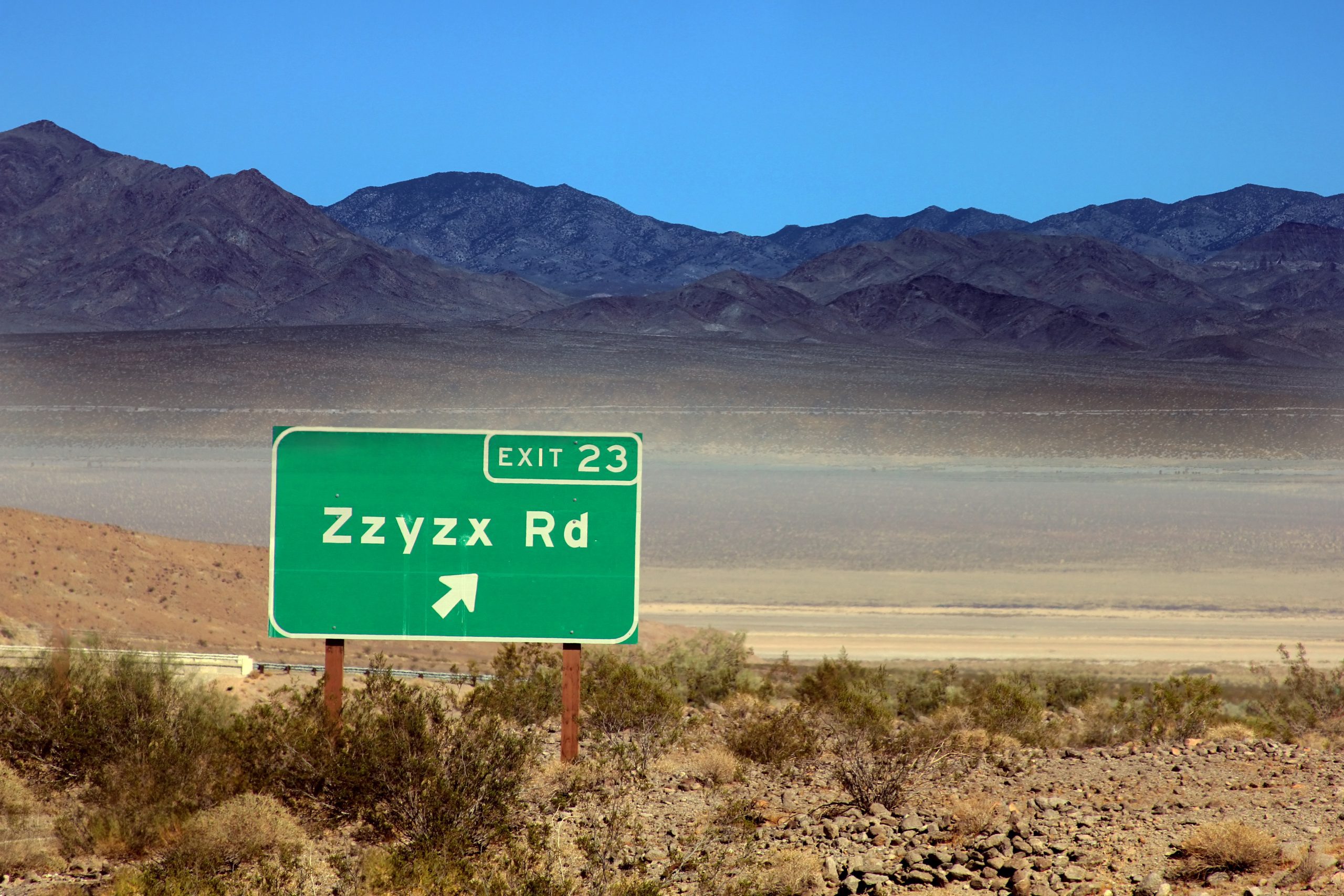 Road sign indicating Exit 23 for Zzyzx Road with a desert landscape and mountains in the background.
