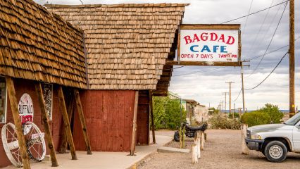 Exterior view of the Bagdad Cafe, featuring a rustic wooden building with a large sign, an old bicycle, and a parked car under a cloudy sky.