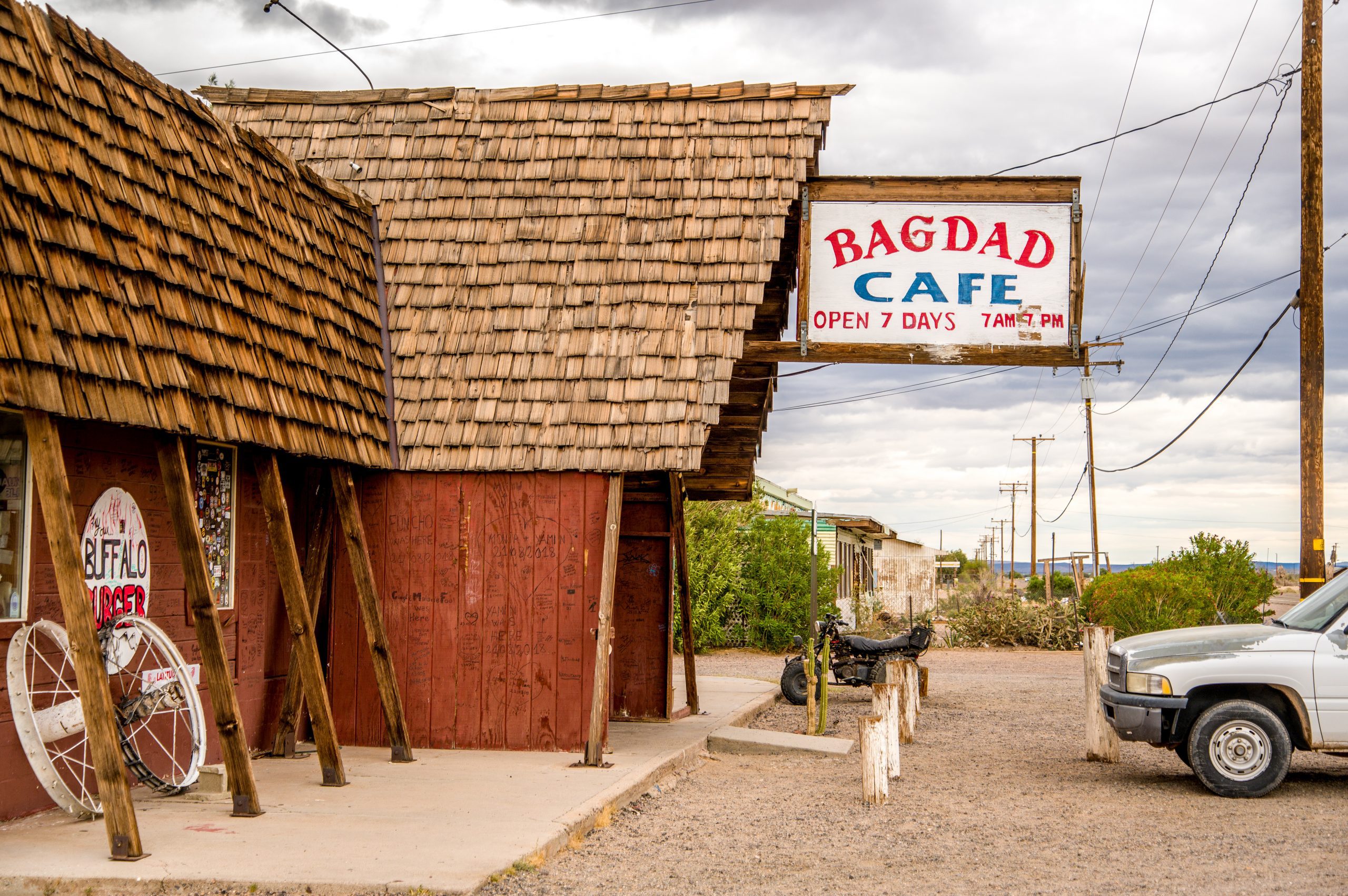 Exterior view of the Bagdad Cafe, featuring a rustic wooden building with a large sign, an old bicycle, and a parked car under a cloudy sky.