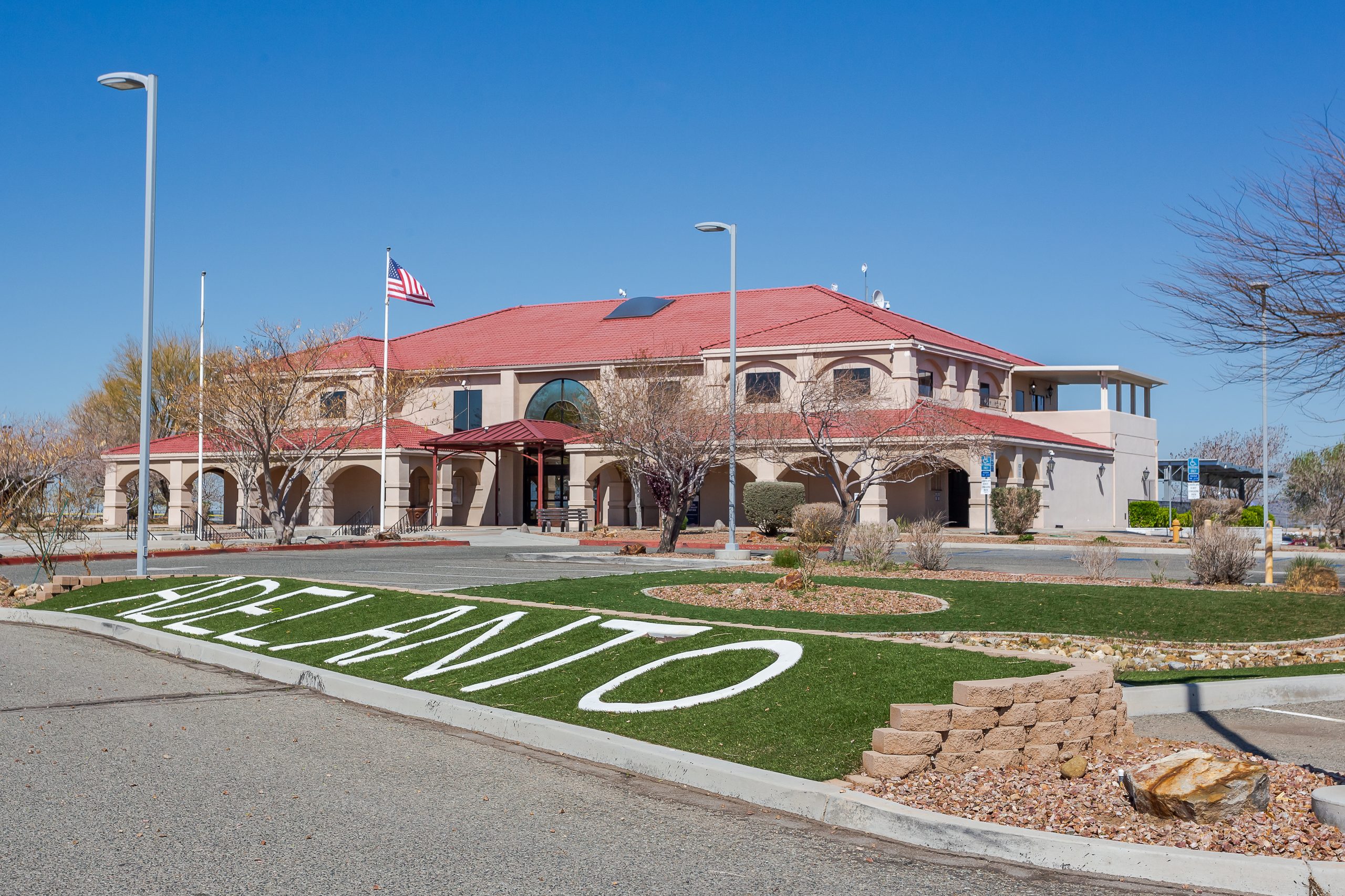A city hall building surrounded by trees, light posts and grass with the word "Adelanto" painted across it.