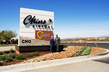 Two men stand beside the Chino Airport monument sign featuring the San Bernardino County logo and CNO airport code, with landscaped grounds, a clear blue sky and mountains in the background.