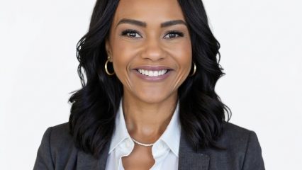 Smiling woman in a gray blazer and white blouse, wearing hoop earrings and a necklace, against a plain light background.