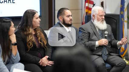 Panel discussion with four people seated; a man in a suit speaks into a microphone, others listen, with a video play button.