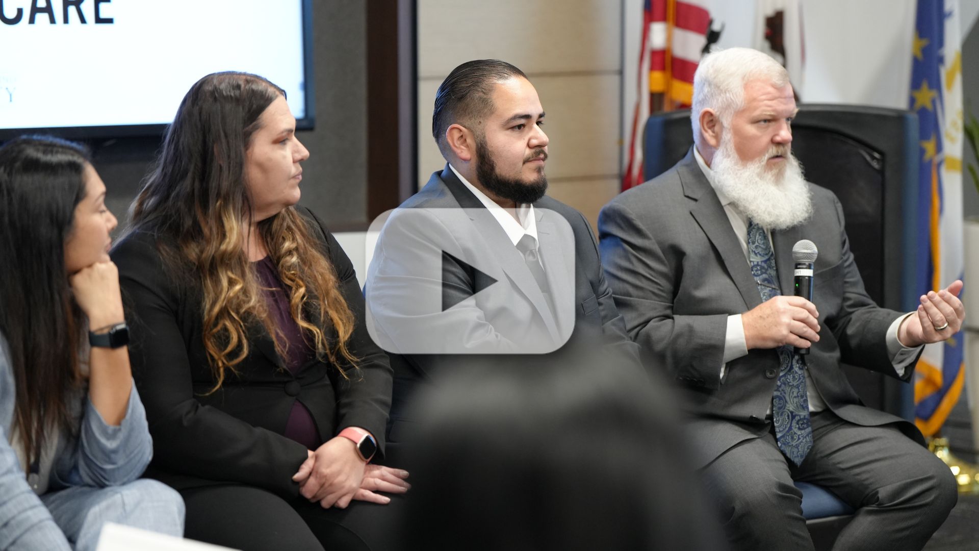 Panel discussion with four people seated; a man in a suit speaks into a microphone, others listen, with a video play button.
