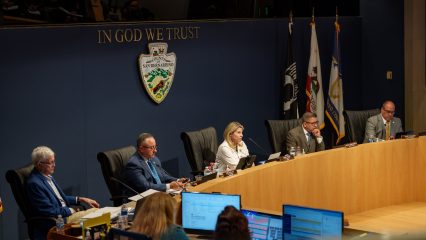 Officials seated at a dais beneath “In God We Trust” and the San Bernardino County seal.