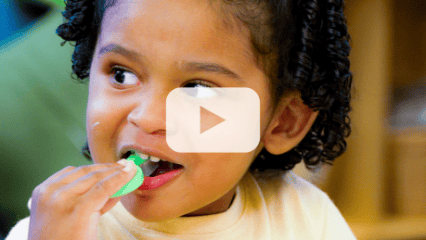 Young child brushes their teeth with a green toothbrush, with a video play button overlay.