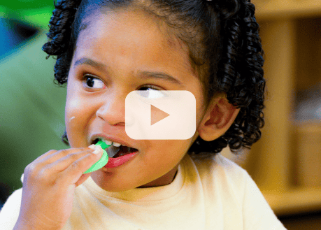 Young child brushes their teeth with a green toothbrush, with a video play button overlay.