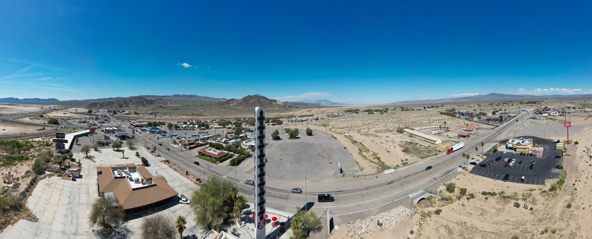 A panoramic view of the World's Tallest Thermometer in Baker. The image also includes various buildings and a roadway with vehicles.