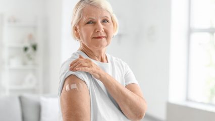 Older woman in a white shirt shows a small bandage on her upper arm after receiving a vaccination.