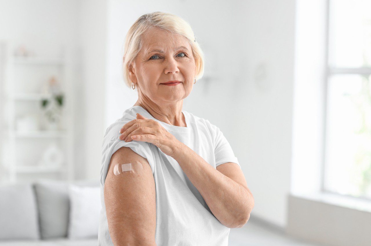Older woman in a white shirt shows a small bandage on her upper arm after receiving a vaccination.