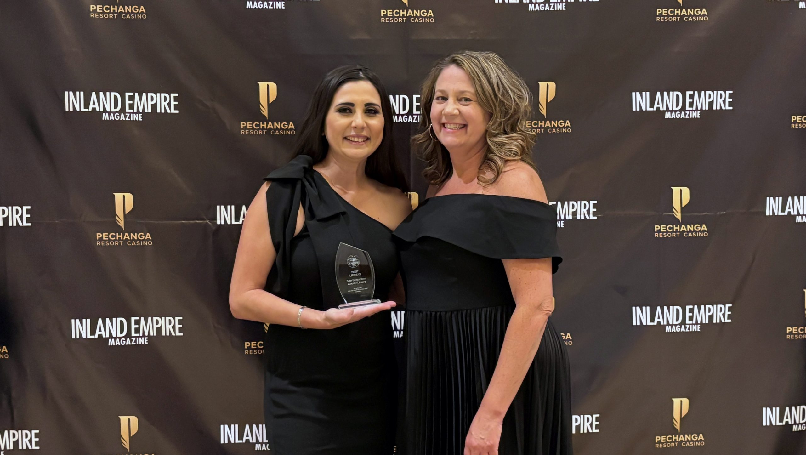 Two women in elegant black dresses pose with an award against a backdrop featuring "Inland Empire Magazine" and "Pechanga Resort Casino" logos.