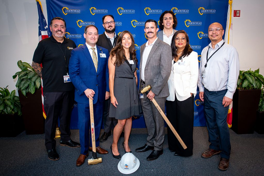 A group of eight smiling people holding hammers and standing in front of a blue backdrop with the San Bernardino County logo at the ARMC Hybrid Bi-Plane Angiography Suite and Operating Room.