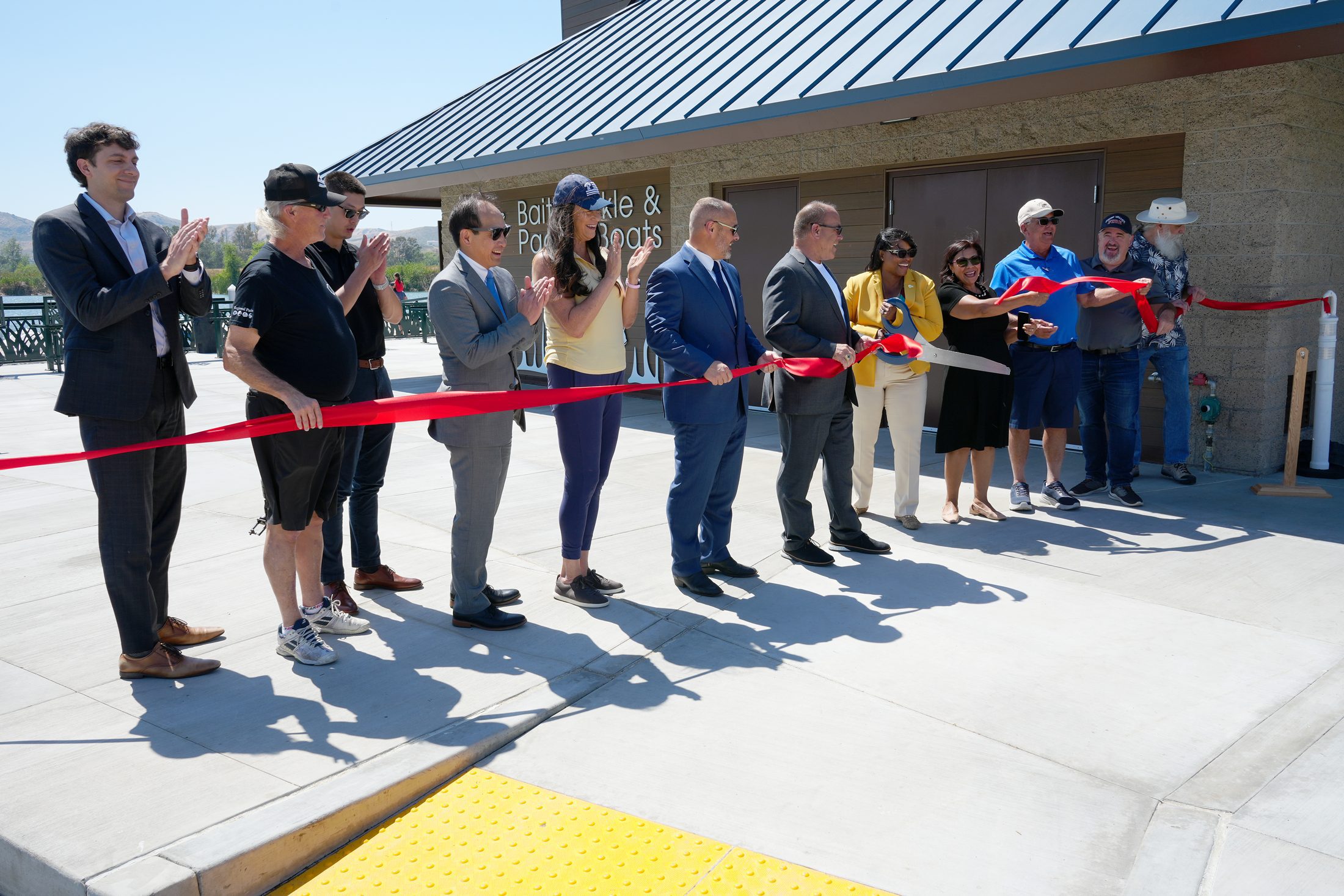 A group of people stand behind a red ribbon with a woman near the center holding a large pair of scissors cutting the ribbon.