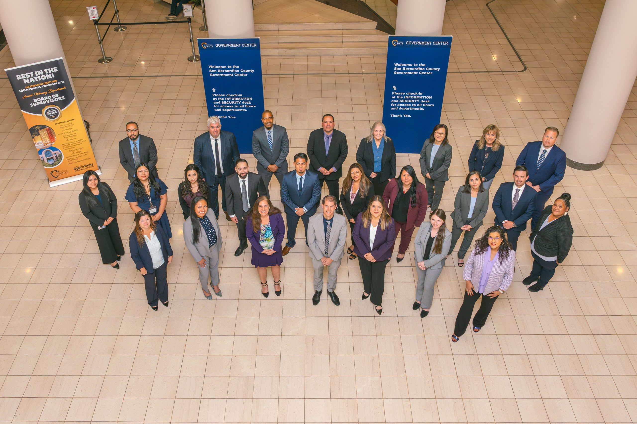 A group of people standing in a lobby in a semi-circle. Two large blue signs behind them display info about the County Government Center.