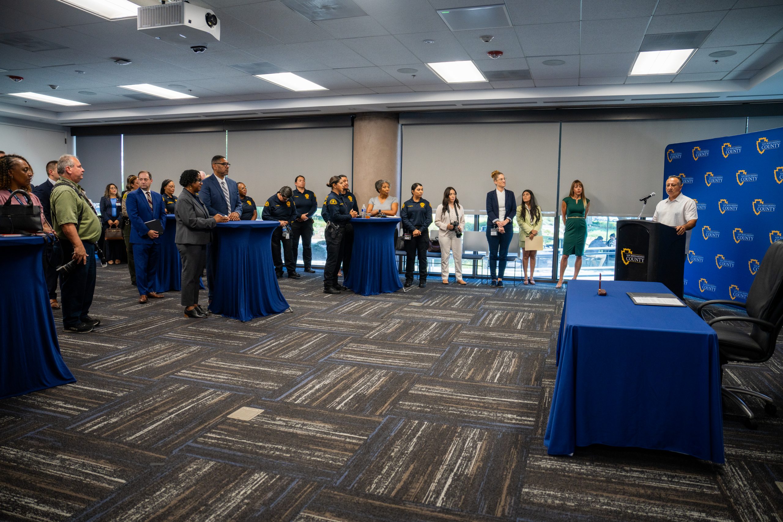 A group of individuals in a conference room listening to a speaker at one of the podiums. The room features gray carpeting and a SB County logo on the backdrop.