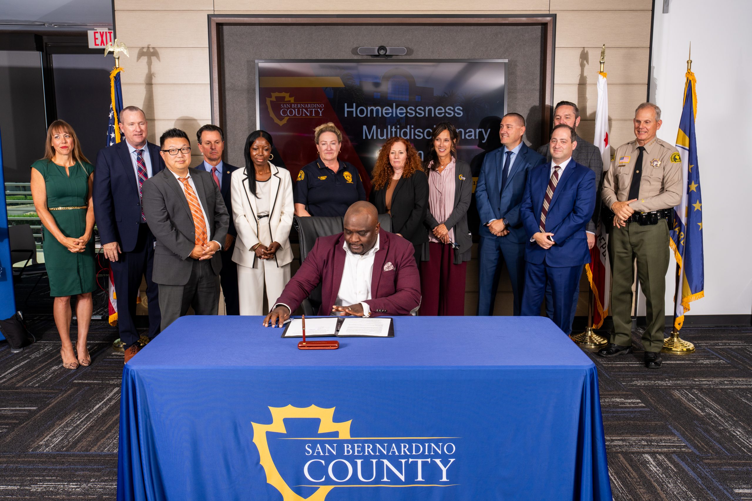 A person signing a document at a table with the San Bernardino County logo, surrounded by a group of eleven individuals standing in a conference room