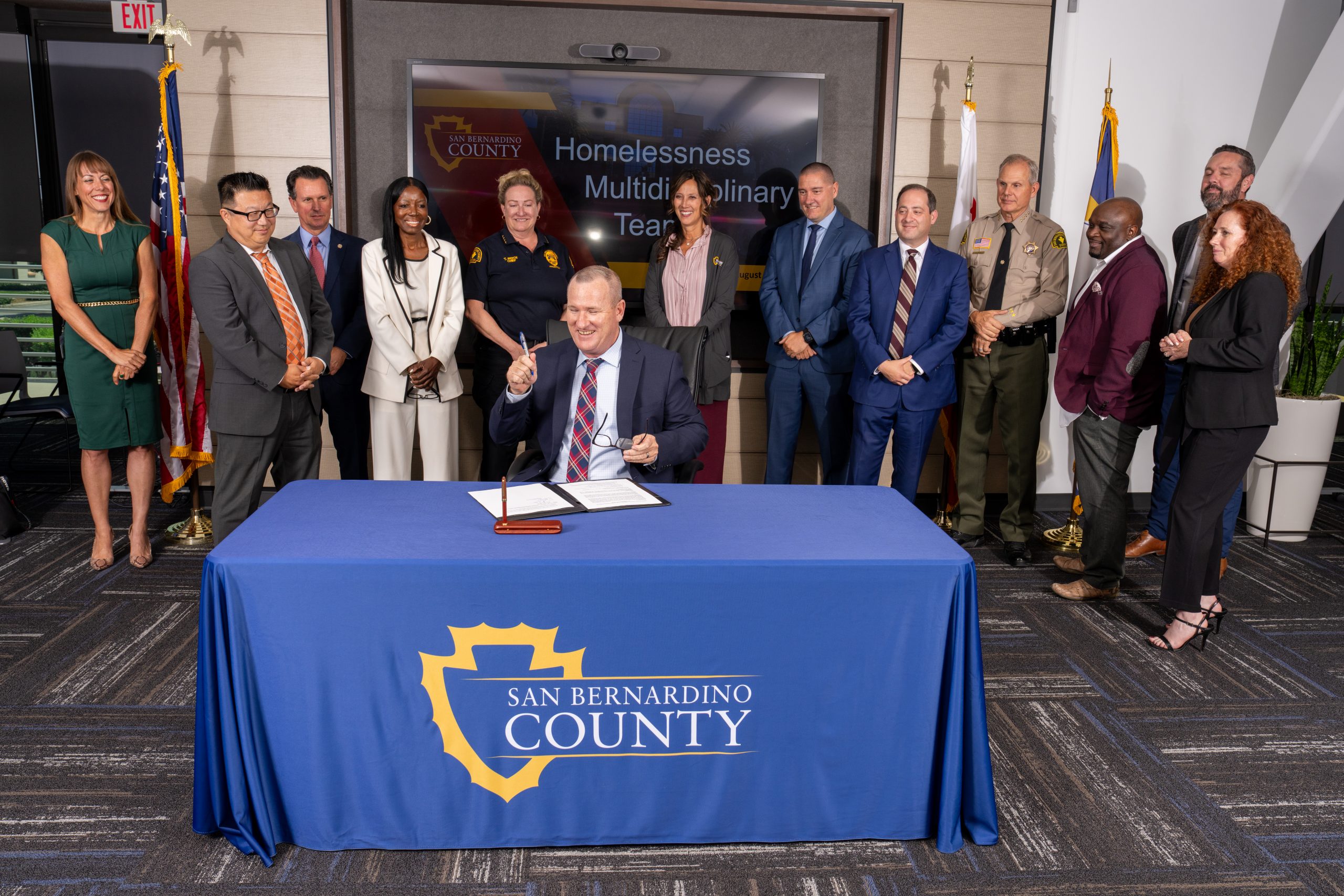 A person signing a document at a table with the San Bernardino County logo, surrounded by a group of individuals standing in a conference room.