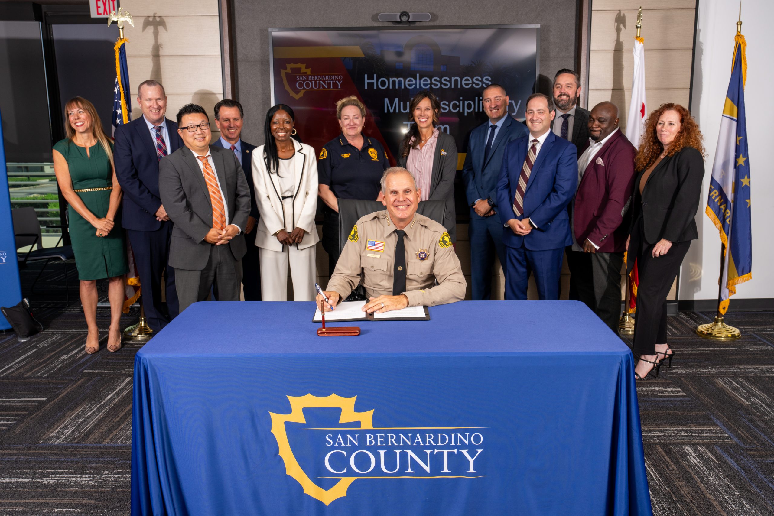 A person signing a document at a table with the San Bernardino County logo, surrounded by a group of individuals standing in a conference room.