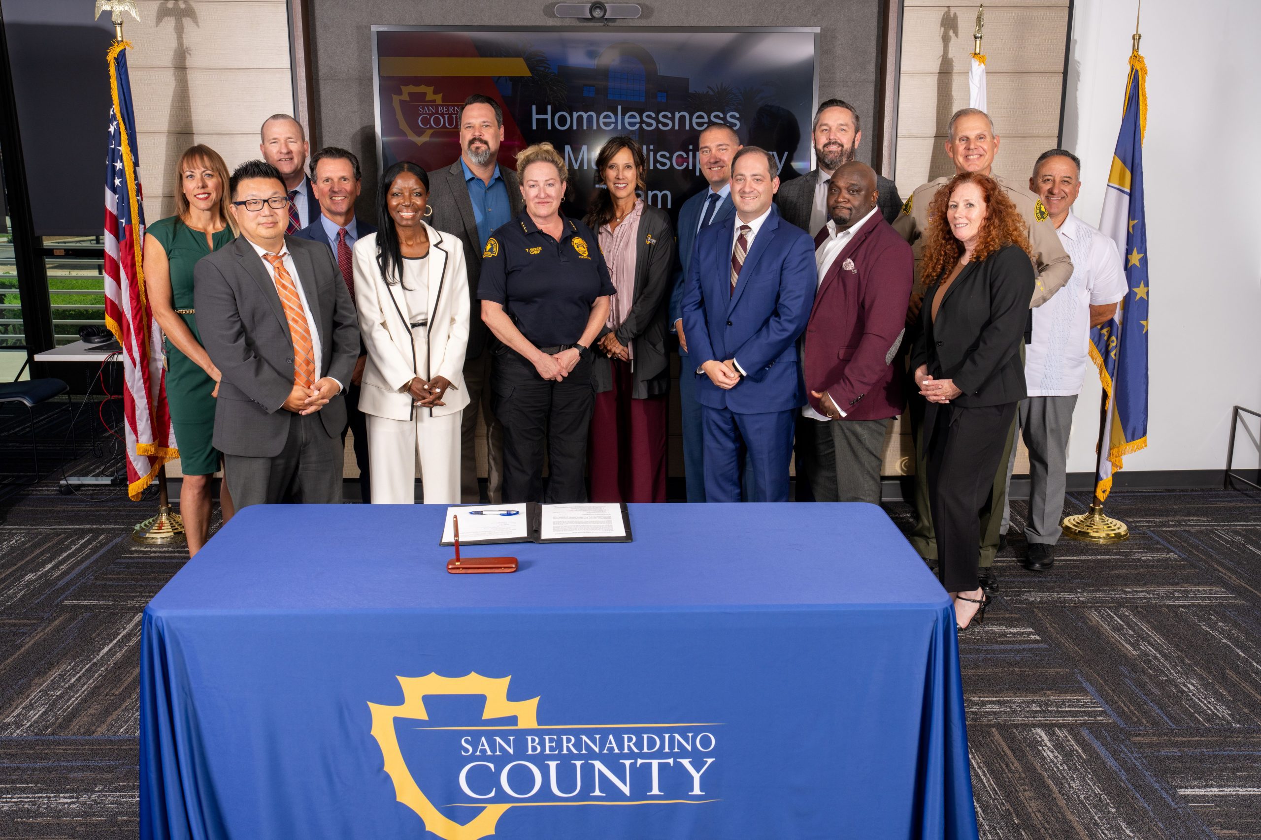 Group of county officials standing behind a table with a sign reading San Bernardino County.