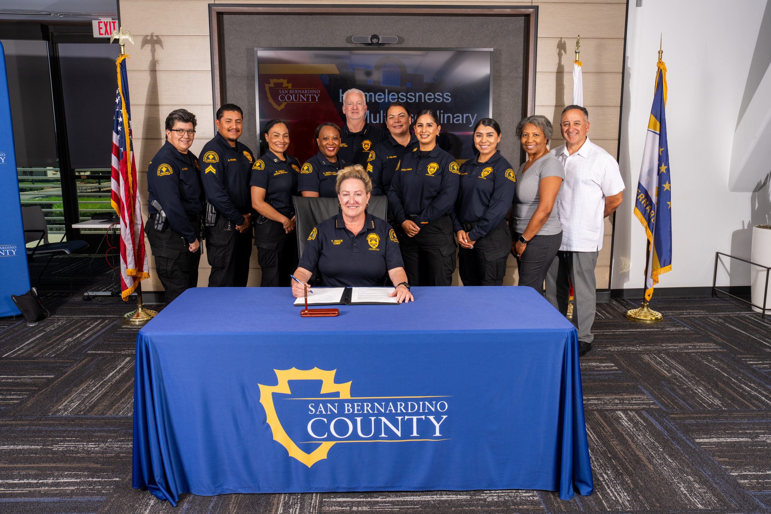 A person signing a document at a table with the San Bernardino County logo, surrounded by a group of individuals standing in a conference room.