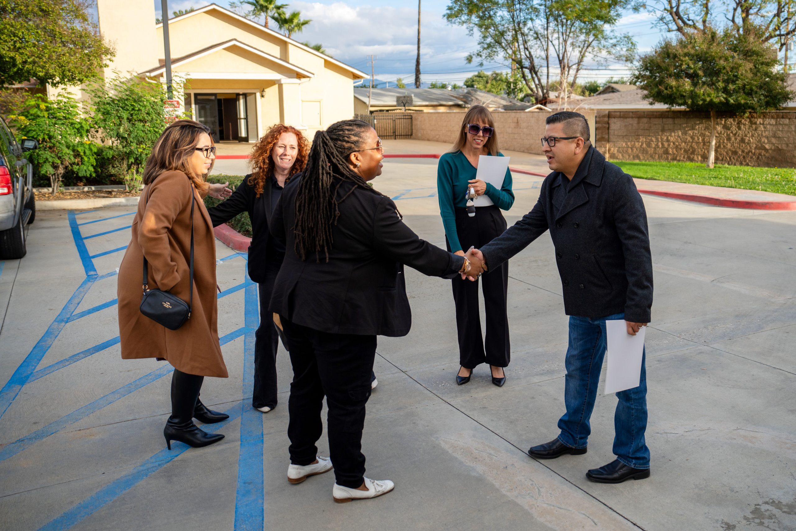 Business professionals shaking hands and interacting in a parking lot outside a building.