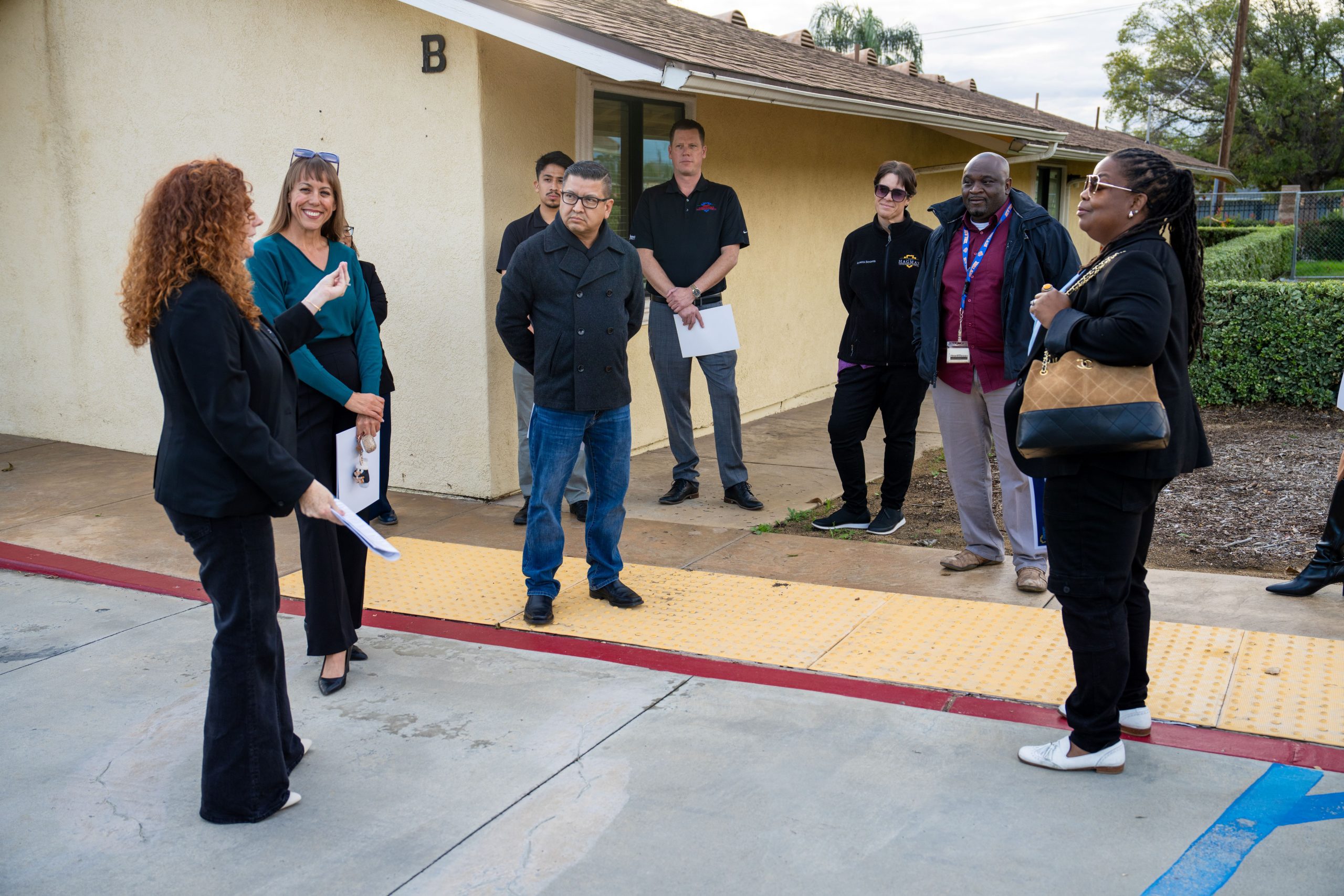 A group of eight adults are engaged in a discussion outside a building marked with the letter 'B'. One person is gesturing with a document, addressing the others who are attentively listening.