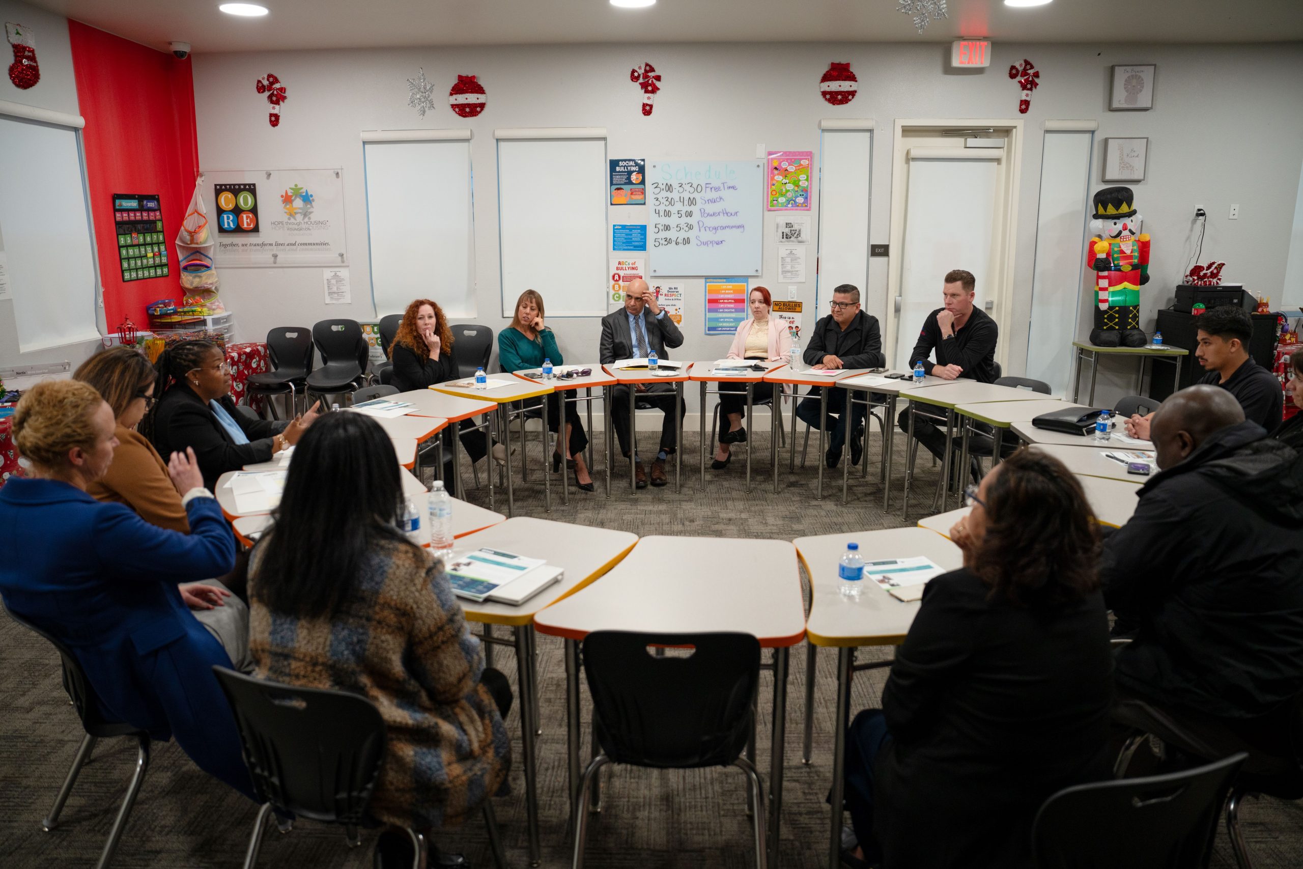 Group of people sitting around a table in a classroom setting, engaged in a discussion. The room is decorated with educational posters and seasonal decorations.