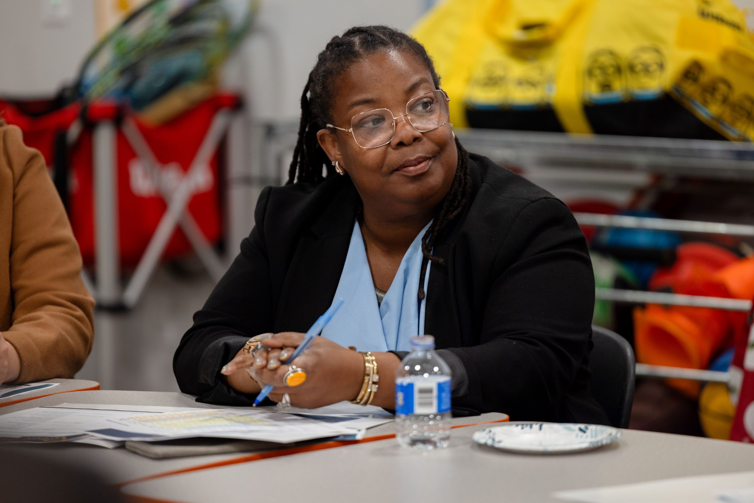 Person sitting at a table during a discussion, wearing a blue shirt and glasses, with various papers and a water bottle in front of them.