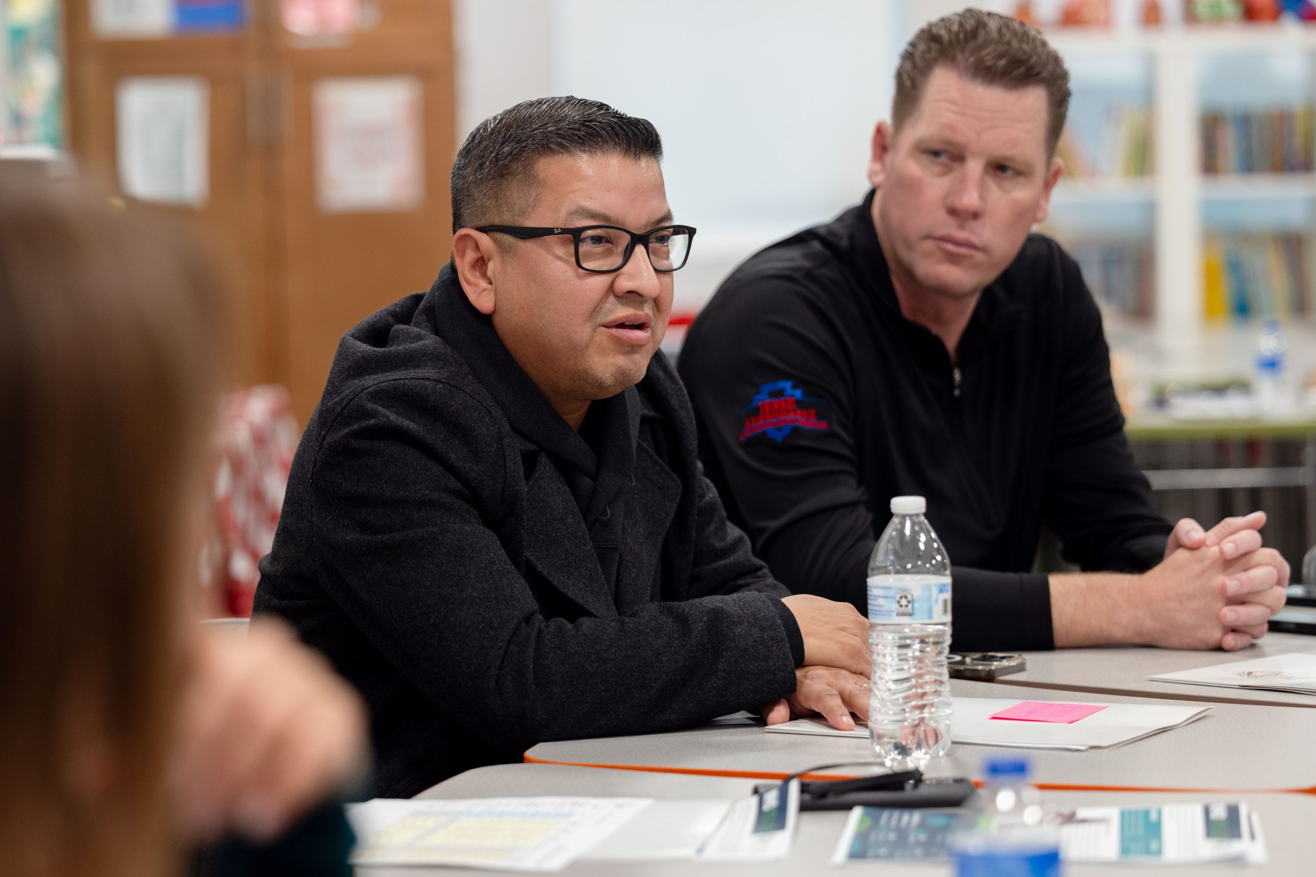 A group of individuals engaged in a discussion at a table with papers and a water bottle, in a room with bookshelves in the background.