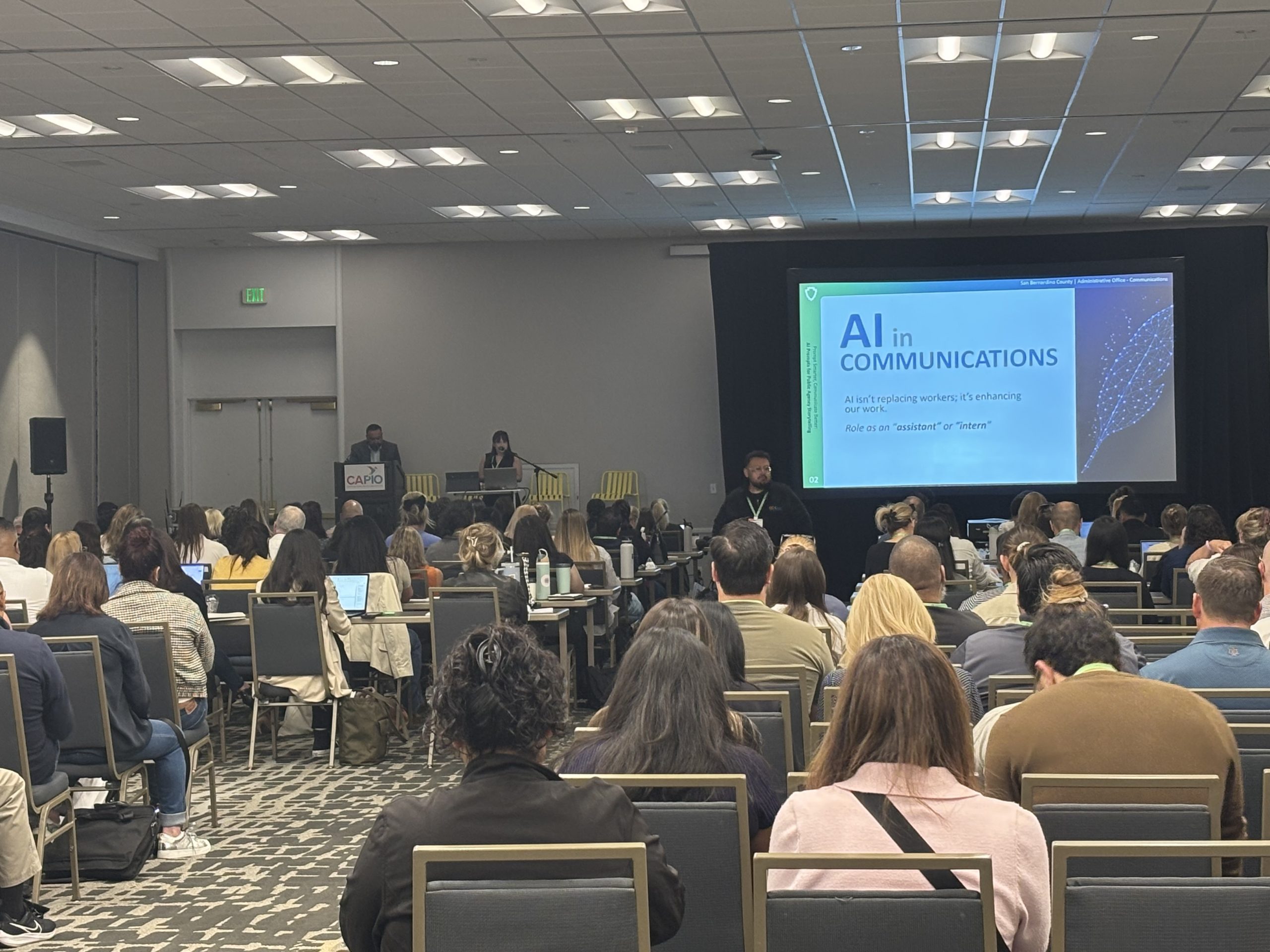 Seated audience in a large conference room with two presenters speaking in the back of the room at a podium and table. Presentation slide reads: “AI in Communications. AI isn’t replacing workers; it’s enhancing our work.”