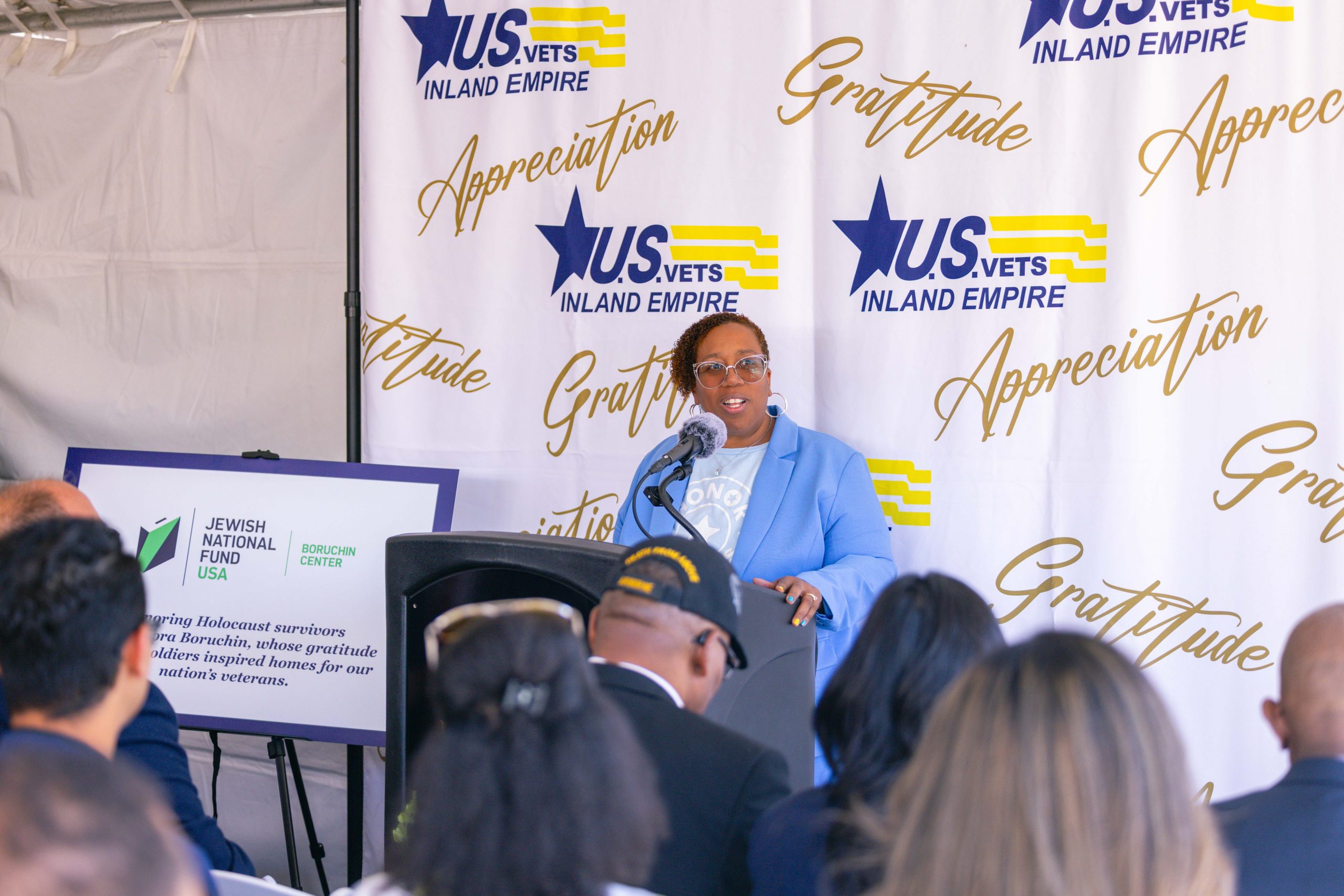 A woman in a light blue suit stands at a podium with a U.S. VETS Inland Empire backdrop, addressing an audience. The setting seems formal, with a U.S. flag to the left and organizational banners surrounding the speaker.