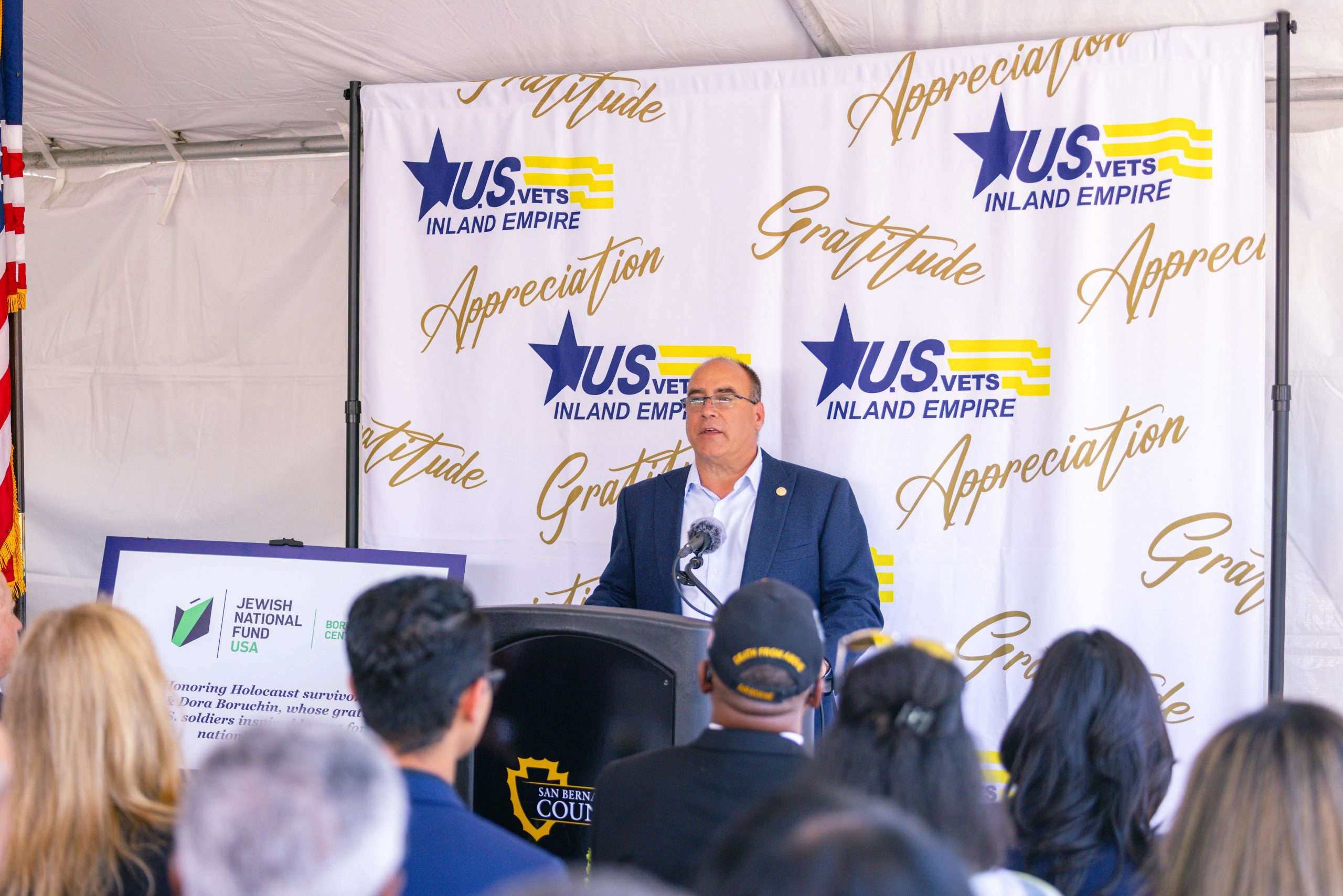 A person stands at a podium with a U.S. VETS Inland Empire backdrop, addressing an audience. The setting seems formal, with a U.S. flag to the left and organizational banners surrounding the speaker.