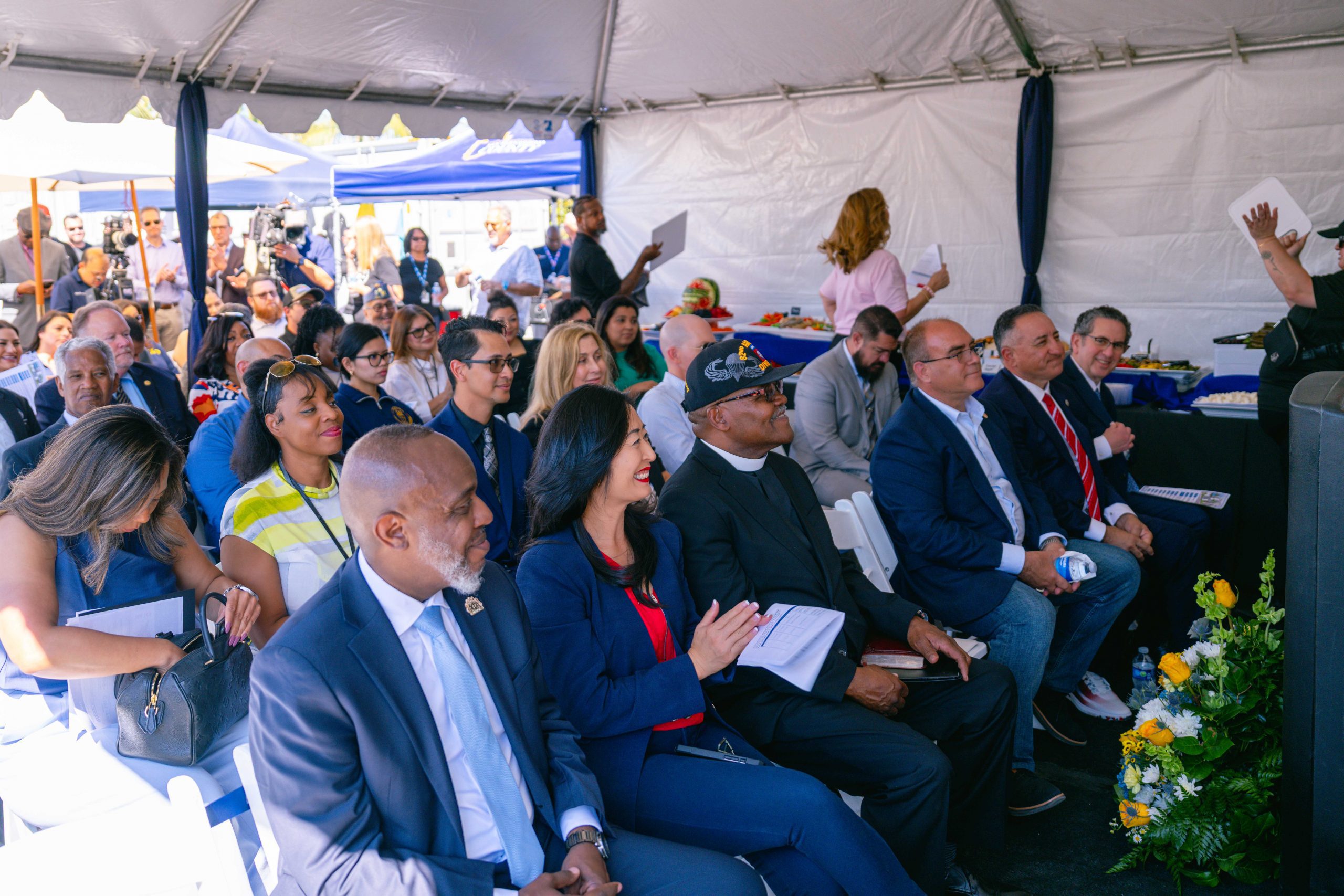 A group of people are seated under a canopy, attending an event. Some are holding papers and others are clapping. There are floral arrangements nearby.