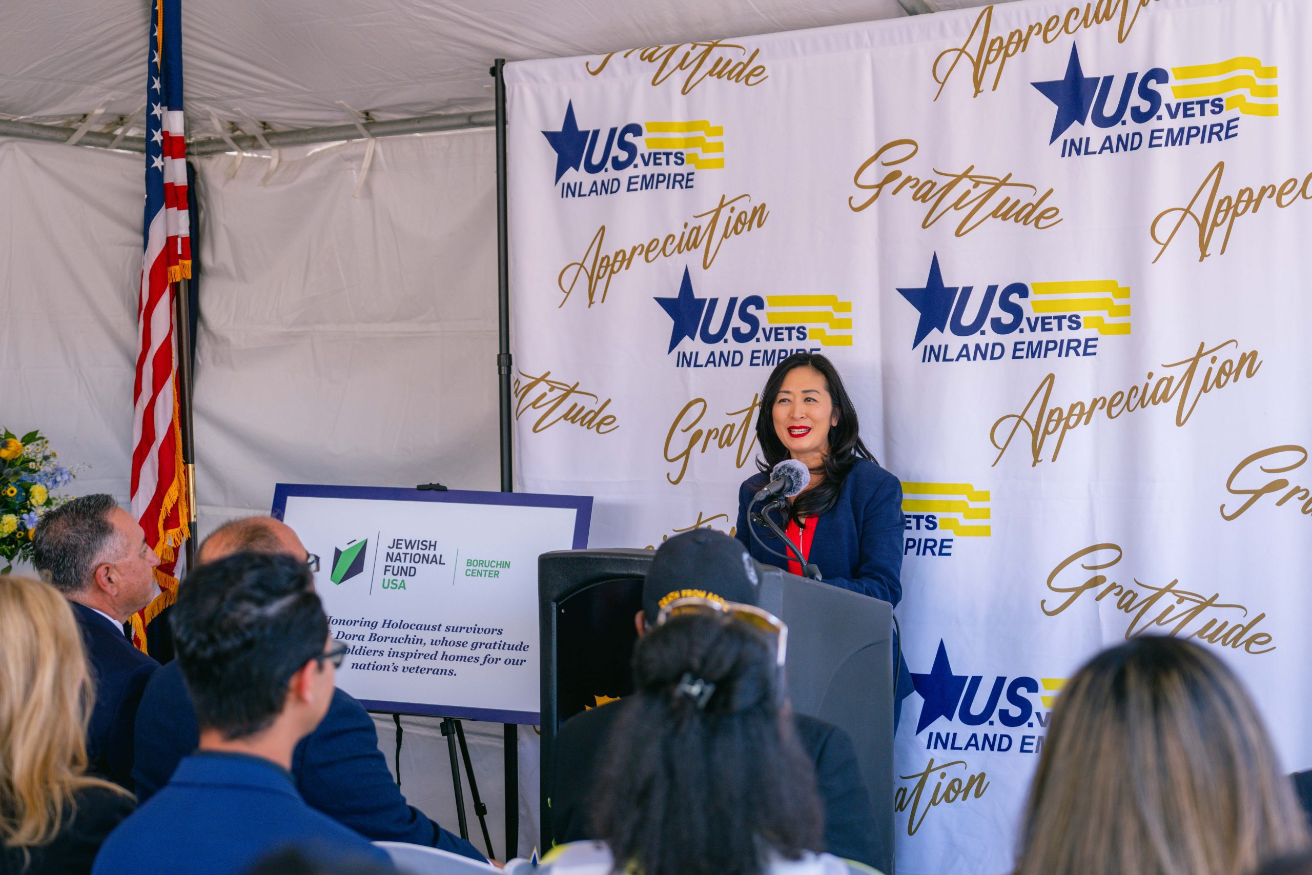 A woman in a navy blue suit stands at a podium with a U.S. VETS Inland Empire backdrop, addressing an audience. The setting seems formal, with a U.S. flag to the left and organizational banners surrounding the speaker.