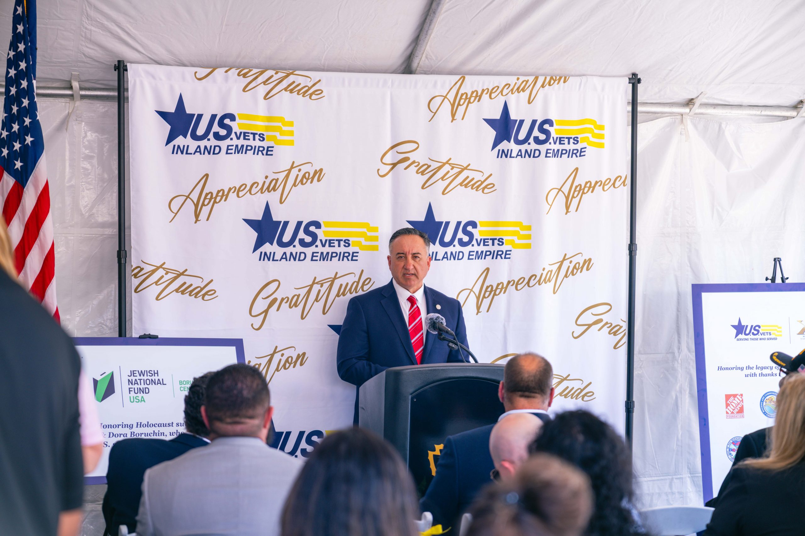A person stands at a podium with a U.S. VETS Inland Empire backdrop, addressing an audience. The setting seems formal, with a U.S. flag to the left and organizational banners surrounding the speaker.