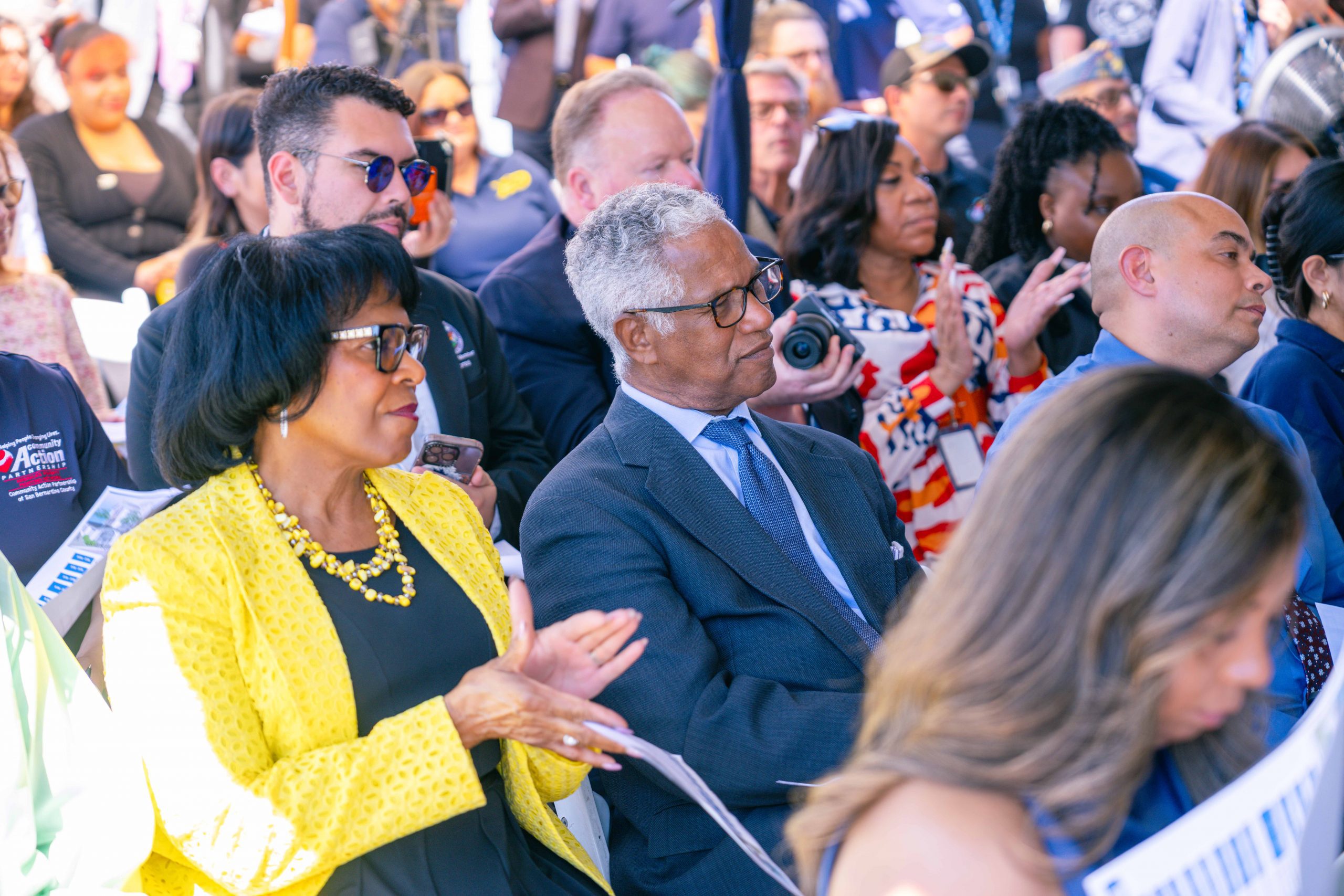 A group of people seated outdoors at an event, including individuals in business attire. Some are holding programs or taking photos. The atmosphere appears engaged and attentive.