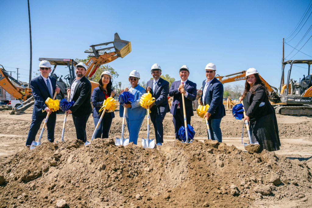 A group of people at a construction site participate in a groundbreaking ceremony. They are dressed in business attire and wearing hard hats.