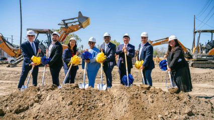 A group of people at a construction site participate in a groundbreaking ceremony. They are dressed in business attire and wearing hard hats.