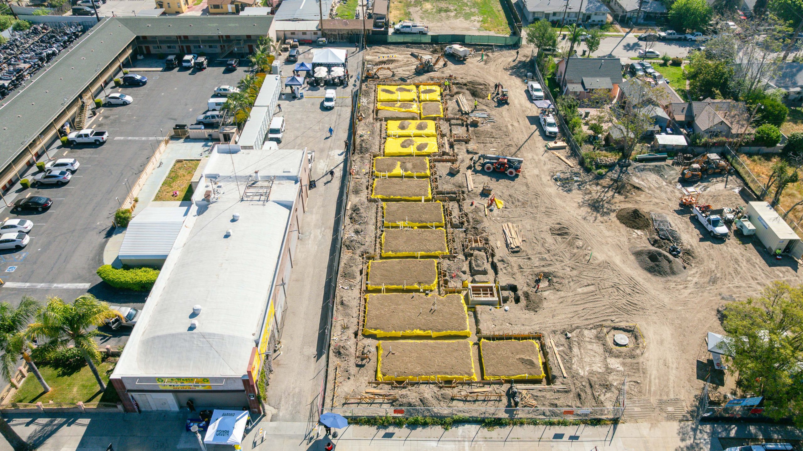 Aerial view of an urban construction site with large, organized rectangles of soil bordered by yellow barriers. Adjacent buildings and parked vehicles are visible. The surrounding area includes streets and trees.